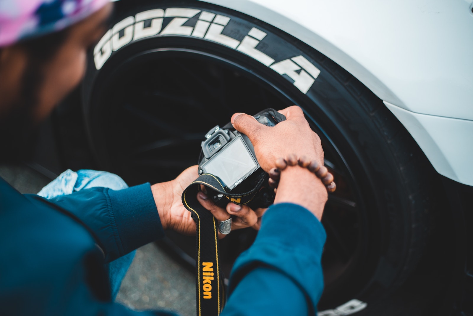 From above back view of anonymous bearded photographer in casual wear taking photo of contemporary auto wheel on digital camera with display and strap on pavement of city street