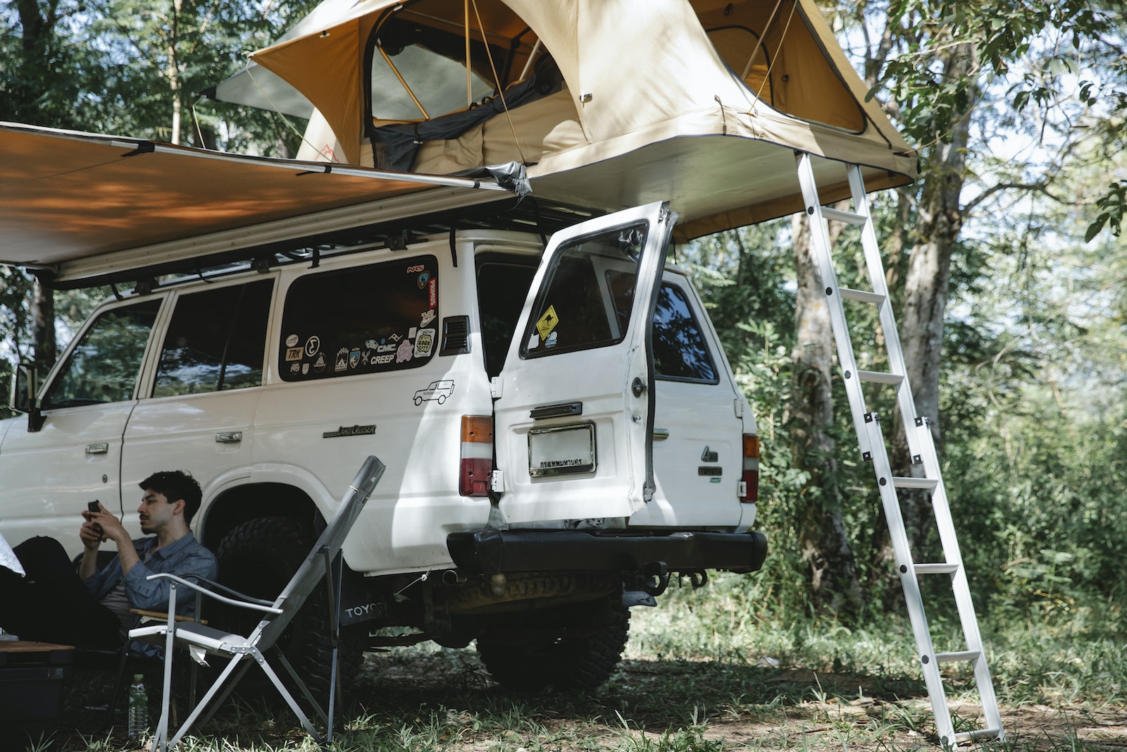 Man sitting under awning placed near offroader with tent on roof in forest